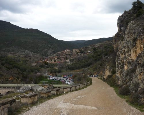 Camino de tierra en descenso en el monte, con una vista panorámica de un pueblo a lo lejos.