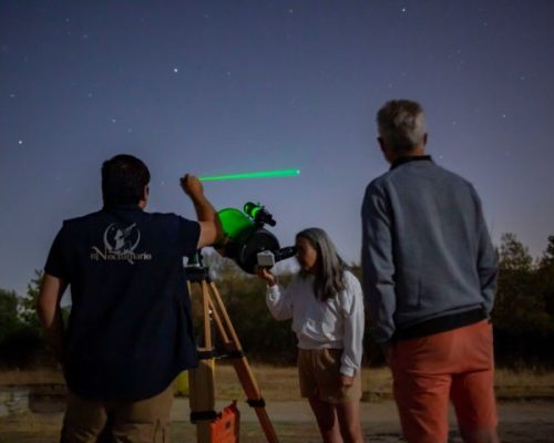 Grupo de personas realizando observación astronómica con telescopio y puntero láser bajo un cielo estrellado.